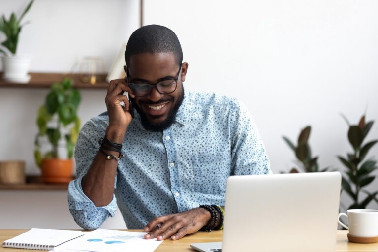 A smiling Black man with buzzed hair and facial hair, wearing a light blue button-up shirt, black-rimmed glasses and bracelets, calls someone on a cell phone while looking at a statistics and has a white laptop open in front of him.