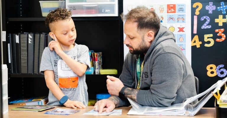 A male teacher helps a male student while sitting a table in a classroom