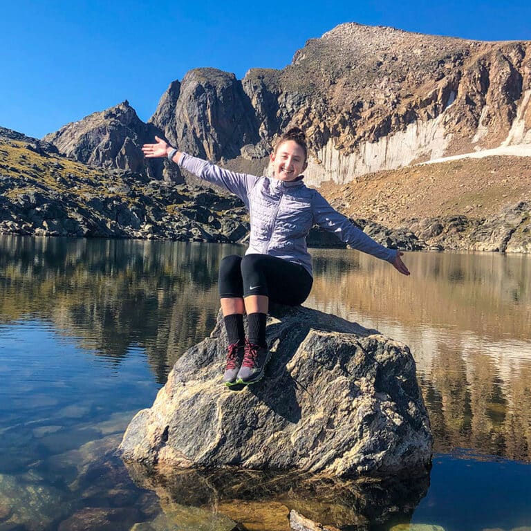 Emily Brixey, outside, sitting on a boulder in a lake surrounded by mountains