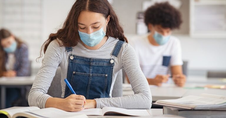 with long curly hair student in overalls and long-sleeve shirt and medical mask writing on a testing booklet at a desk with other students in the background