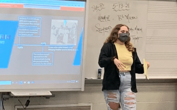 teenager with long curly, auburn hair in a mask, yellow shirt, black cardigan, and ripped blue jeans speaking in fornt of a projected powerpoint on mascot imagery
