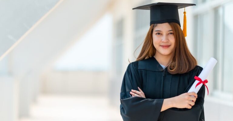 Portrait of a young woman with straight, long auburn hair smiling at the camera, holding a diploma and in a graduation uniform