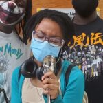 a teenaged Black girl with locks wearing a blue mask and cardigan sings at a microphone