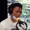 A teenaged Black boy with locks, wearing a white t-shirt and headphones speaks at a microphone.