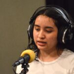 A high school girl with dark hair and brown skin wears headphones and sits in front of a microphone speaking