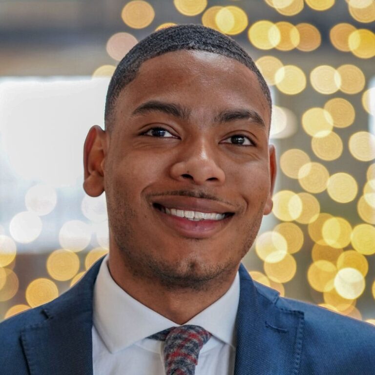 Photo portrait of Deion A. Jordan, a young Black man with a buzz cut in a suit and tie with lights in the background