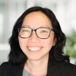 A woman with dark hair wearing dark blazer and glasses smiles at the camera