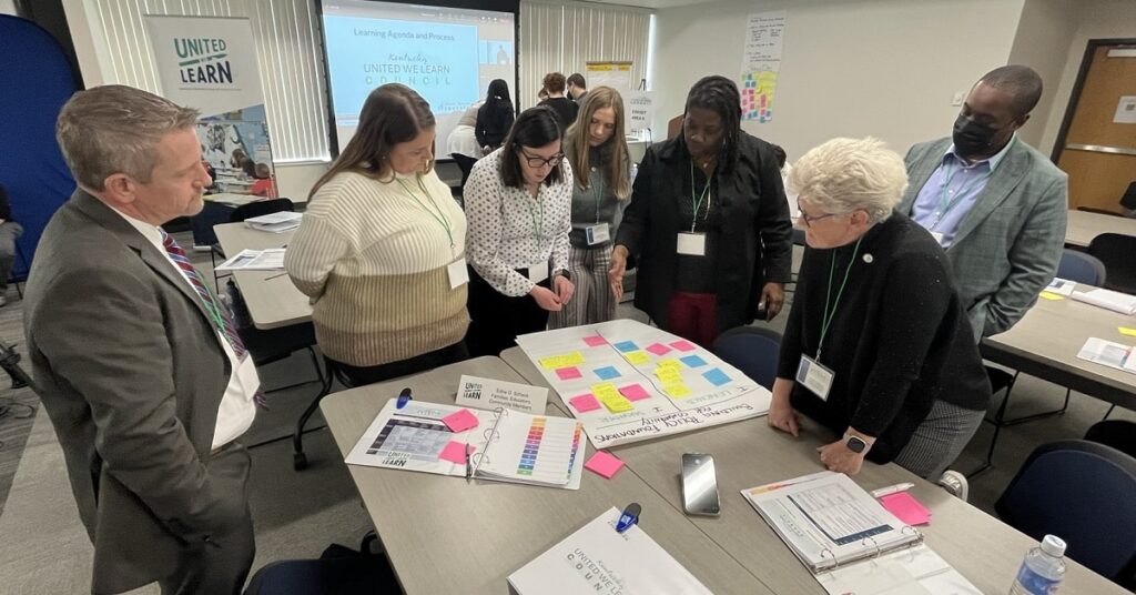 a group of people convened at a table talking over a board with sticky notes