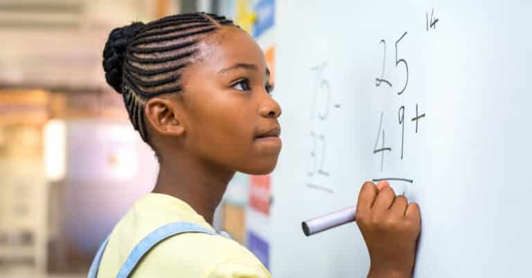 a middle-school aged Black girl with cornrows pulled back into a bun in bib overalls and yellow shirt at a white board solving a math problem