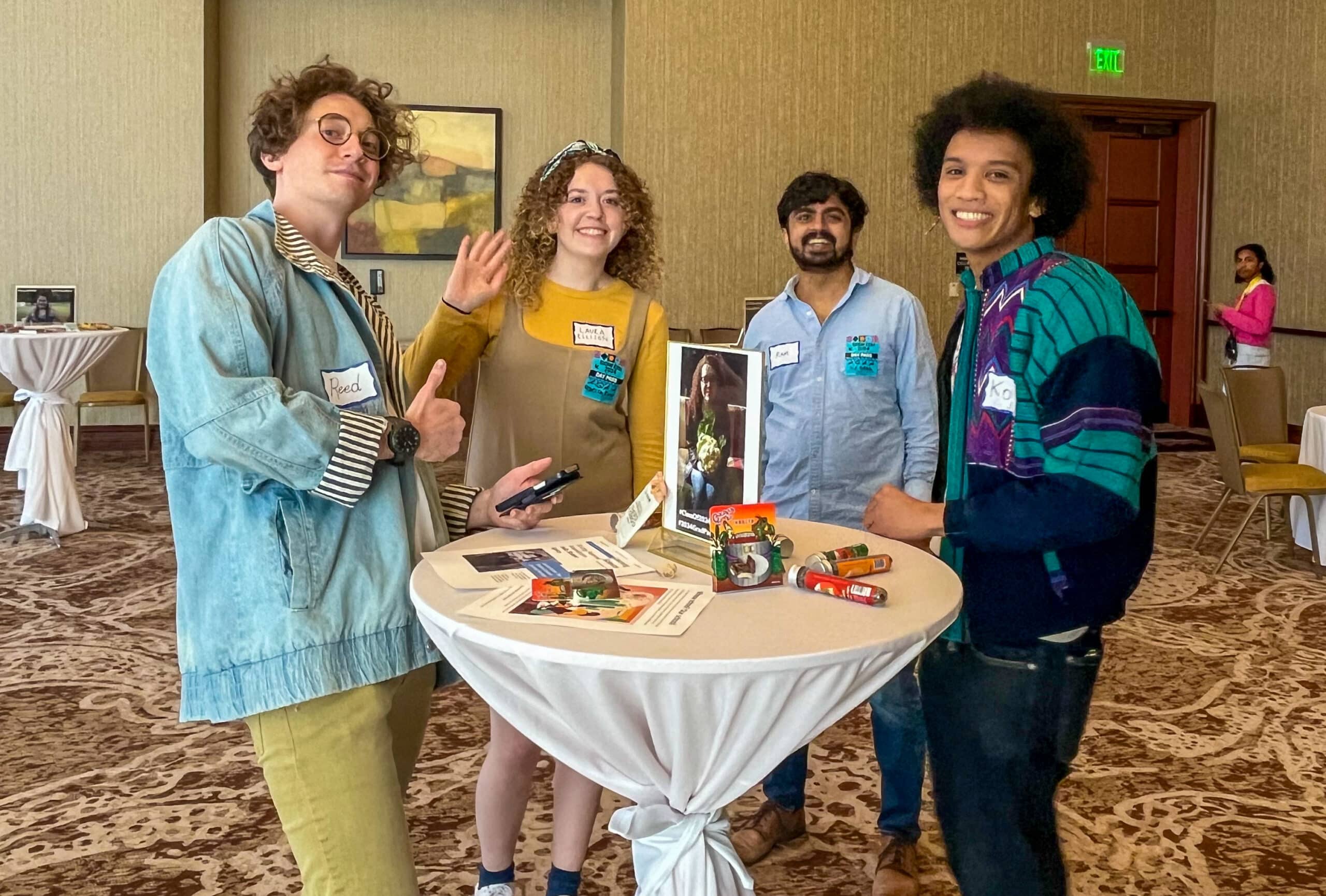 Young adults standing around a table, smiling and waving at the camera