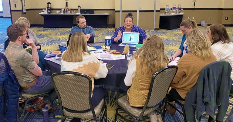 A group of North Dakota educators sit around a table talking with papers and computers in front of them. One is speaking animatedly.
