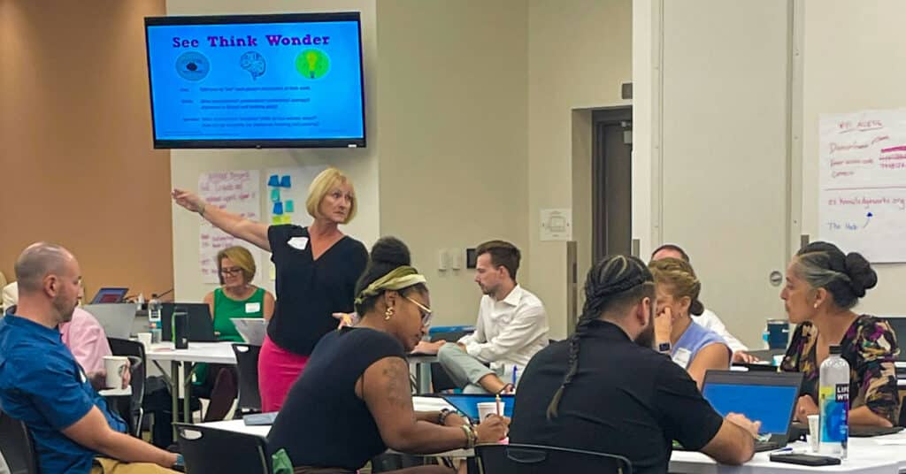 Educators convened at tables with one standing, guiding them through instructions