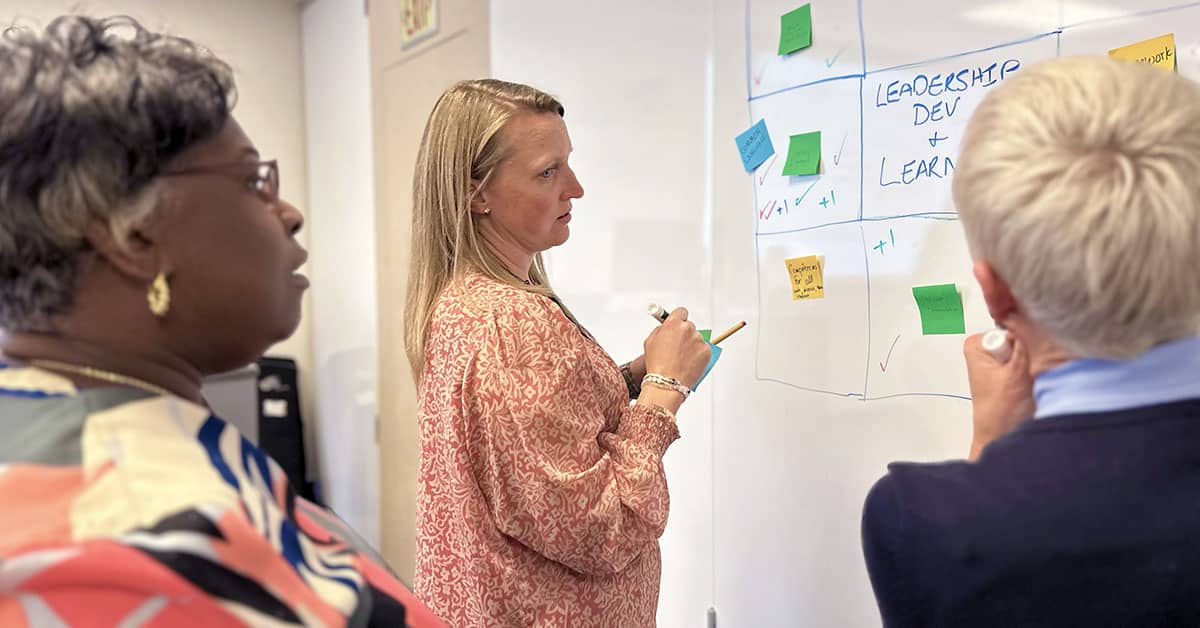 Three women of varying ages and race examine a chart on the wall with sticky notes attached