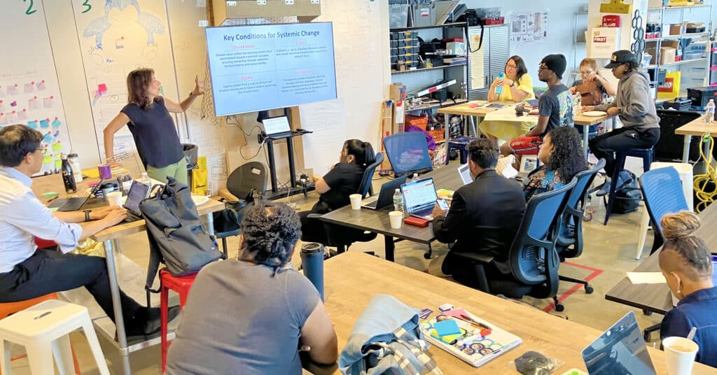 Educators and learners gathered in a room with the speaker at the front pointing attention to the TV screen next to her