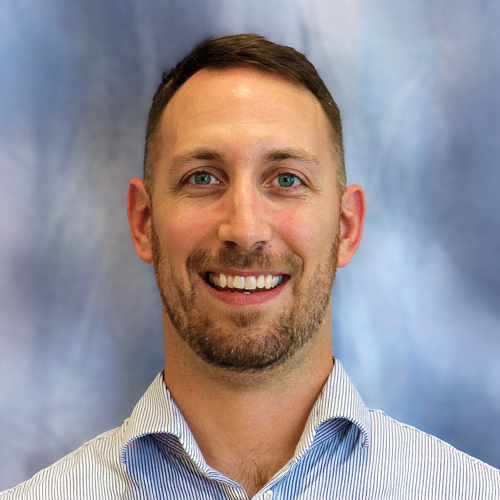 Headshot of Alex Polumbo, a man with with short facial hair and brown hair, wearing a light blue button-up shirt.