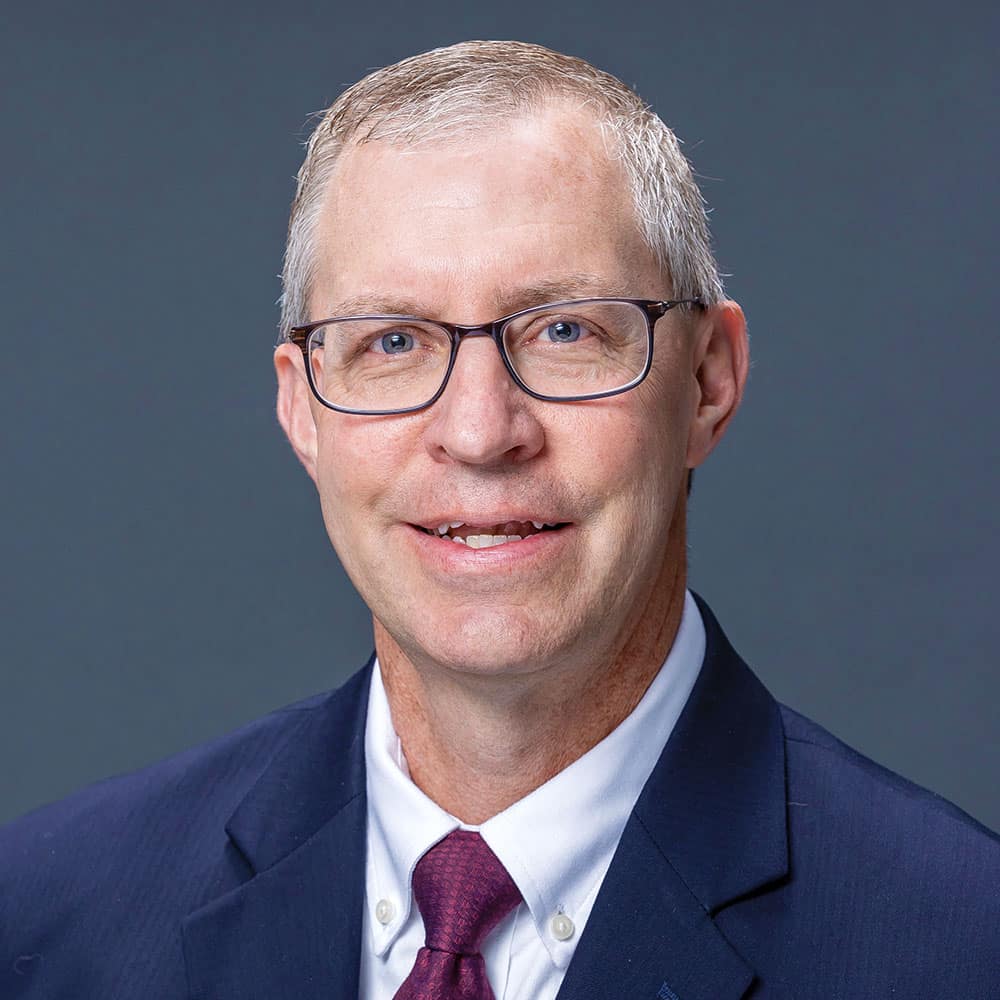 headshot of Joel Schleicher, a clean-shaven White man with short white hair, wearing a navy suit jacket, crimson tie and white button-up