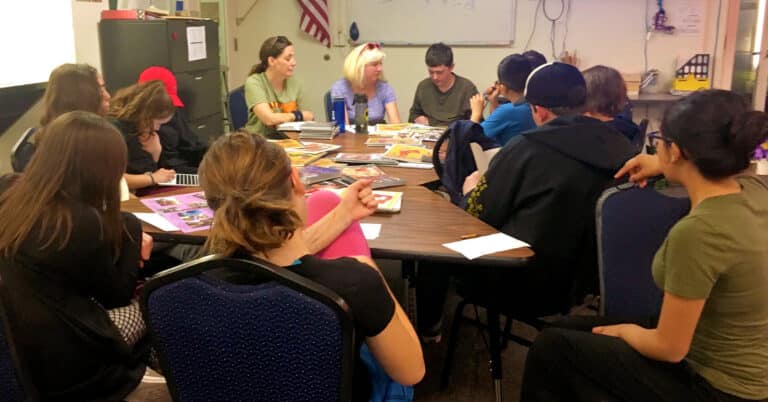 A large group of teens and educators sits around tables pushed together, pay attention to a teen talking