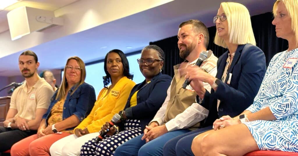 A group of men and women of varying ages and races in business attire sit in stools on a stage. One woman is speaking to the audience as the others focus on her.