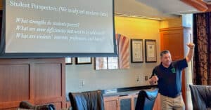 A man animatedly presents to an audience in front of a screen that reads: “How can we ensure our students are equipped to take advantage of career opportunities? Student Perspective: (We analyzed student data) What strengths do students possess? What are some deficiencies that need to be addressed? What are students’ interests, preferences, and habits?”