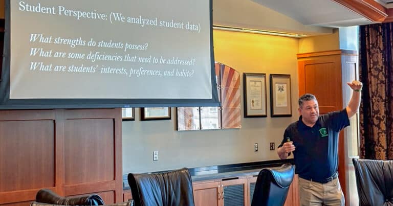 A man animatedly presents to an audience in front of a screen that reads: “How can we ensure our students are equipped to take advantage of career opportunities? Student Perspective: (We analyzed student data) What strengths do students possess? What are some deficiencies that need to be addressed? What are students’ interests, preferences, and habits?”