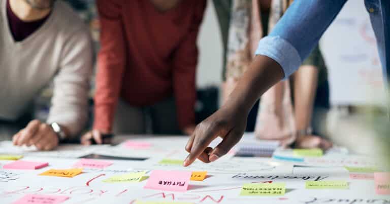 among blurry forms of people leaning over a table of sticky notes, an arm clearly points out to one sticky note