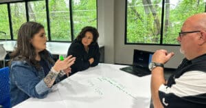 Three educators converse at a table with a green wooded area outside