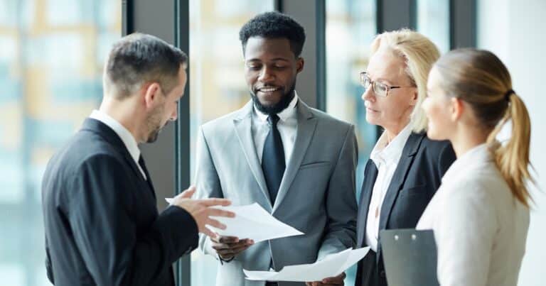 A group of four people gather to examine and discuss what's on the content of a paper one is holding
