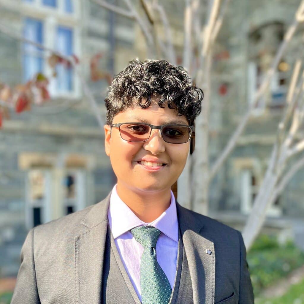 headshot of Vallu Pendyala, a nonbinary brown-skinned person with short curly dark hair, smiling at the camera, wearing sunglasses, a light gray suit, a green tie and a lavender button-up