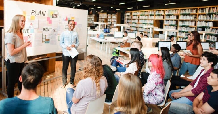 A diverse group in a library setting, with a two people in front of a white board facilitating or presenting to the group