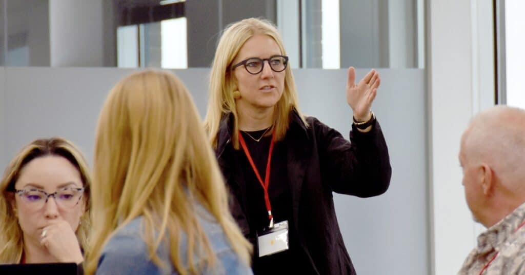 A blonde white man stsands in front of a group sitting at tables with her hand out, animatedly talking