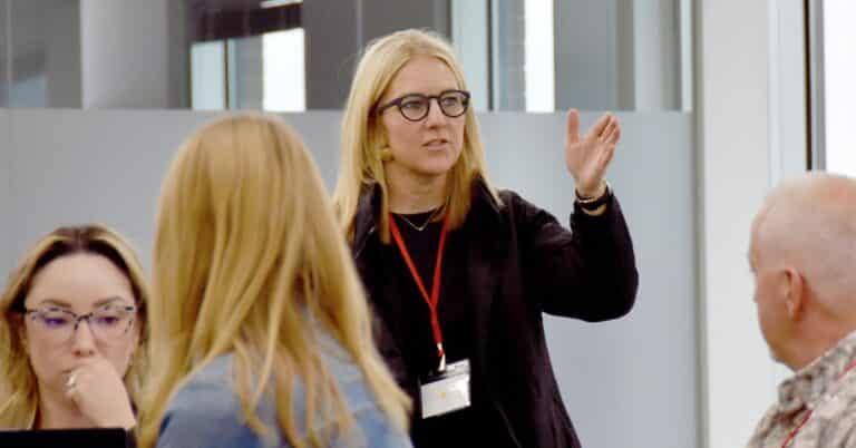 A blonde white man stsands in front of a group sitting at tables with her hand out, animatedly talking