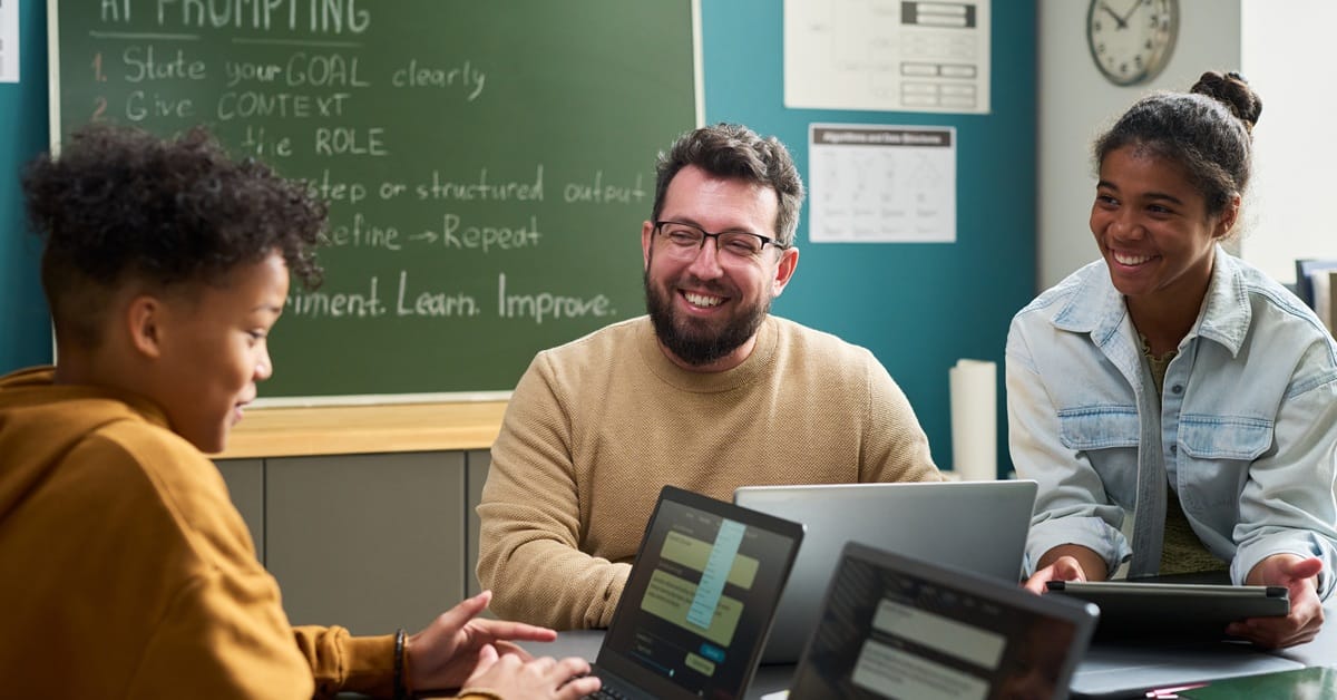 Caucasian man smiling and interacting with Black teenage girl and Black teenage boy using laptops in classroom setting, chalkboard with AI prompting instructions visible in background