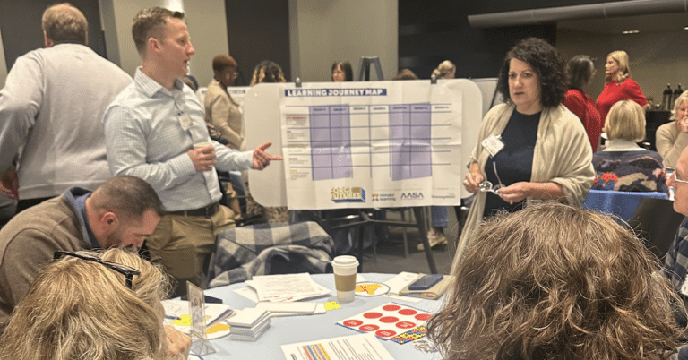 A group of adults gather in a large event space around a table, talking to each other in front of a chart on an eisel