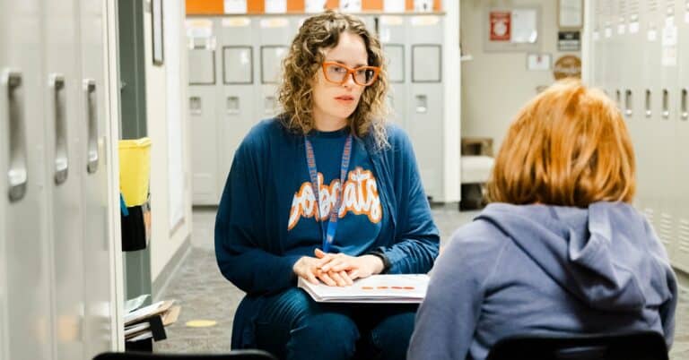 In a hallway surrounded by lockers, a blonde female educator, wearing red glasses and a blue Bobcats sweatshirt, sits accross from a red-headed learner wearing a hoodie