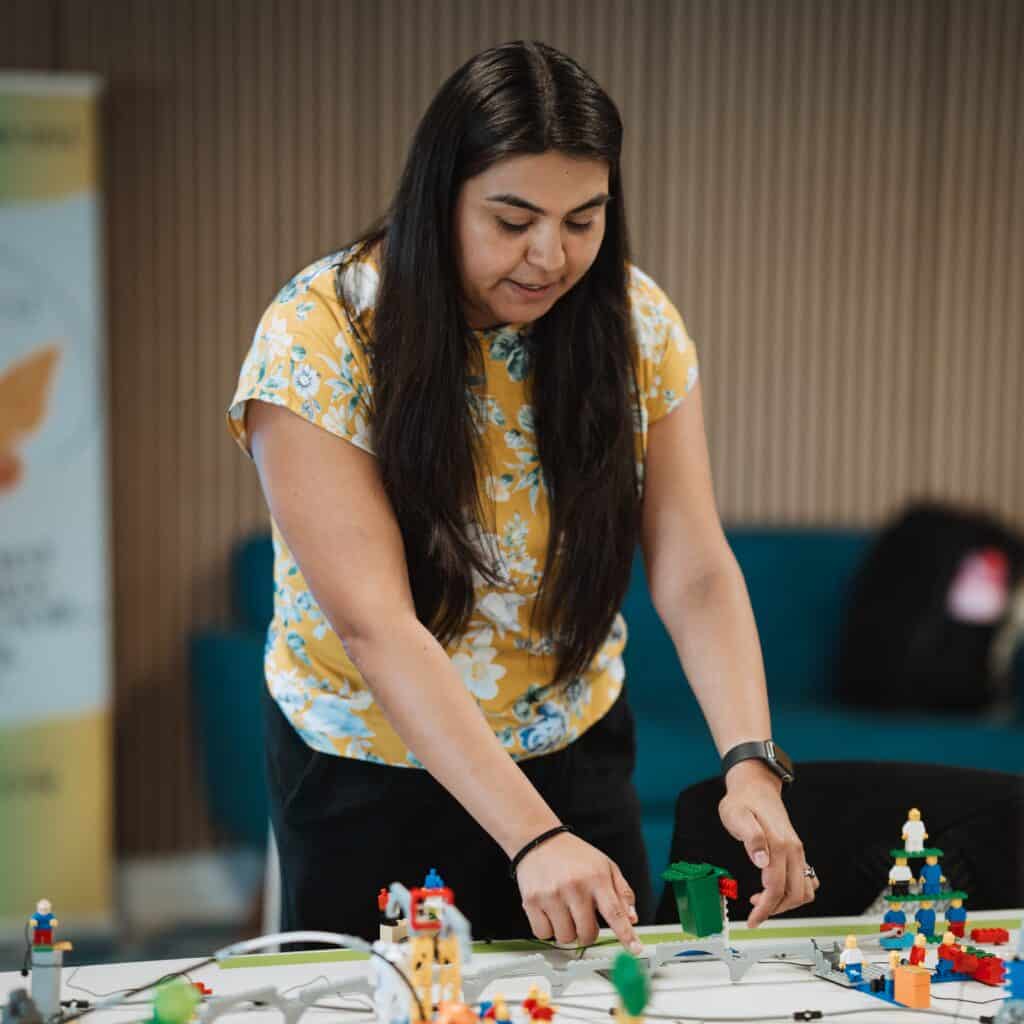A young woman with long, straight dark hair, wearing a blue, gold and white top and black pants is playing with building blocks at a table.
