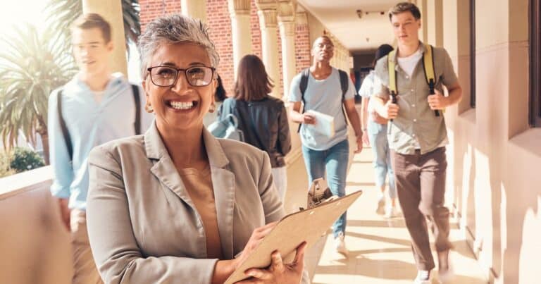 As teens walk in the hallway behind her, smiling for the camera is a brown-skinned woman with silver hair, earing and glasses in a gray blazer and tan shirt under it with a clipboard in her hands