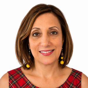A woman with shodulder-length straight honey brown hair smiles at the camera, wearing yellow earrings and a plaid top.